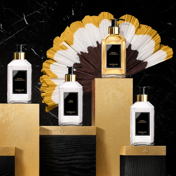 Four elegant Guerlain bottles with black and gold labels, each featuring the GSA dispensing pump with metal-free pathway, are displayed on gold and black pedestals. Behind them, a decorative fan adds flair against the black marble background.