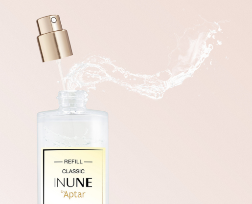 A perfume bottle labeled INUNE by Aptar with liquid spraying out in a curved stream features a classic prestige fragrance pump named Classic Inune, set against a light pink background. The fragrance pump is shown above the bottle, detached.