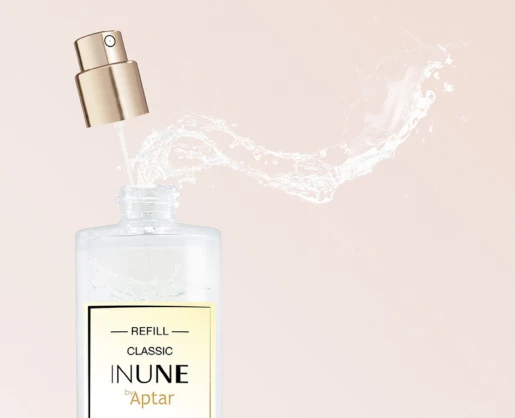 A perfume bottle labeled INUNE by Aptar with liquid spraying out in a curved stream features a classic prestige fragrance pump named Classic Inune, set against a light pink background. The fragrance pump is shown above the bottle, detached.
