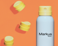 A white spray can labeled Markus by Aptar stands upright on an orange background, featuring a premium twist to lock aerosol design and three yellow aerosol actuators floating nearby.