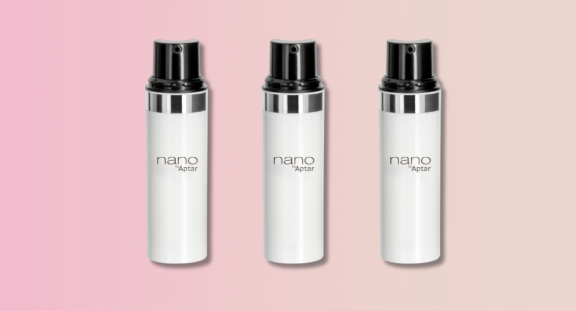 Three white cylindrical bottles with black actuators and a silver metalized band, labeled Nano Aptar, are displayed side by side against a pink-to-beige gradient background, showcasing sleek travel-size airless packaging.