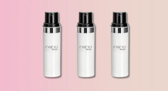 Three white cylindrical bottles with black actuators and a silver metalized band, labeled Nano Aptar, are displayed side by side against a pink-to-beige gradient background, showcasing sleek travel-size airless packaging.