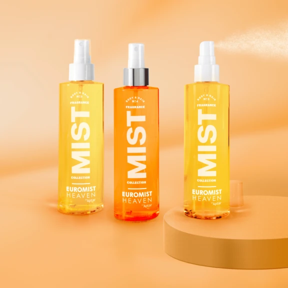 Three spray bottles labeled  EUROMIST HEAVEN stand on a peach-colored surface and background, with one bottle with a silver collar ,and one spraying a fine mist. The bottles are in shades of yellow and orange.