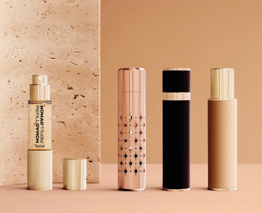 Four elegant cosmetic bottles and tubes, including Refillable Mini Fragrance Packaging, in gold, rose gold, and black tones stand upright on a beige surface before a textured stone background. The containers feature sleek, modern designs.