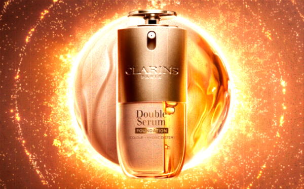 Packaging of Double Serum Foundation of Clarins at the center with a halo of golden light and splashes of serum and foundations formula in the background