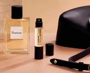 A refillable travel fragrance spray with a logo stating Nomad Refill by Aptar is displayed alongside a full-size fragrance bottle and a purse on a tabletop, showing portable fragrance packaging for on-the-go use.