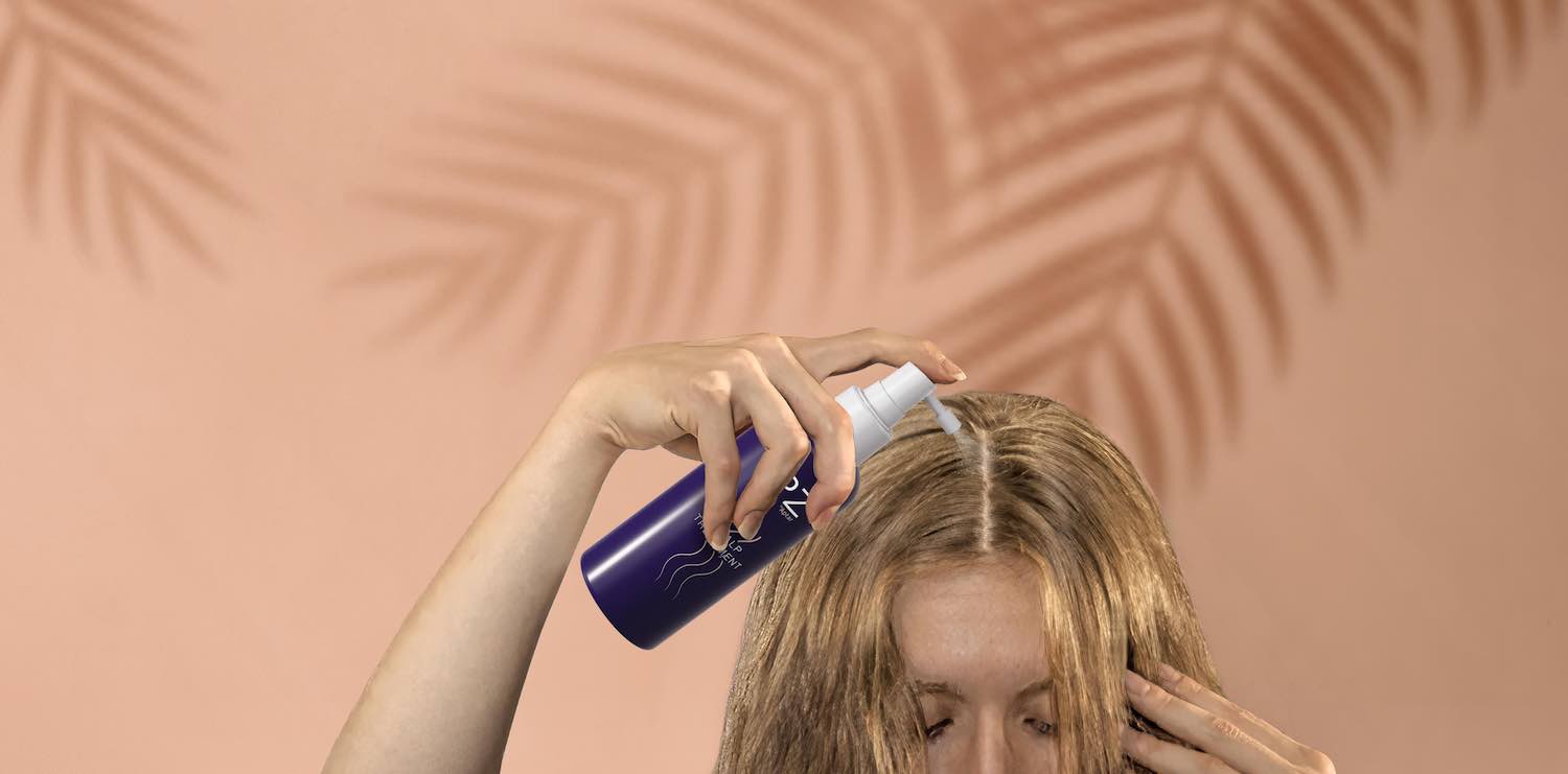 A person sprays a liquid from a blue haircare packaging bottle onto their scalp, parting their blond hair. The background is pink with palm leaf shadows.
