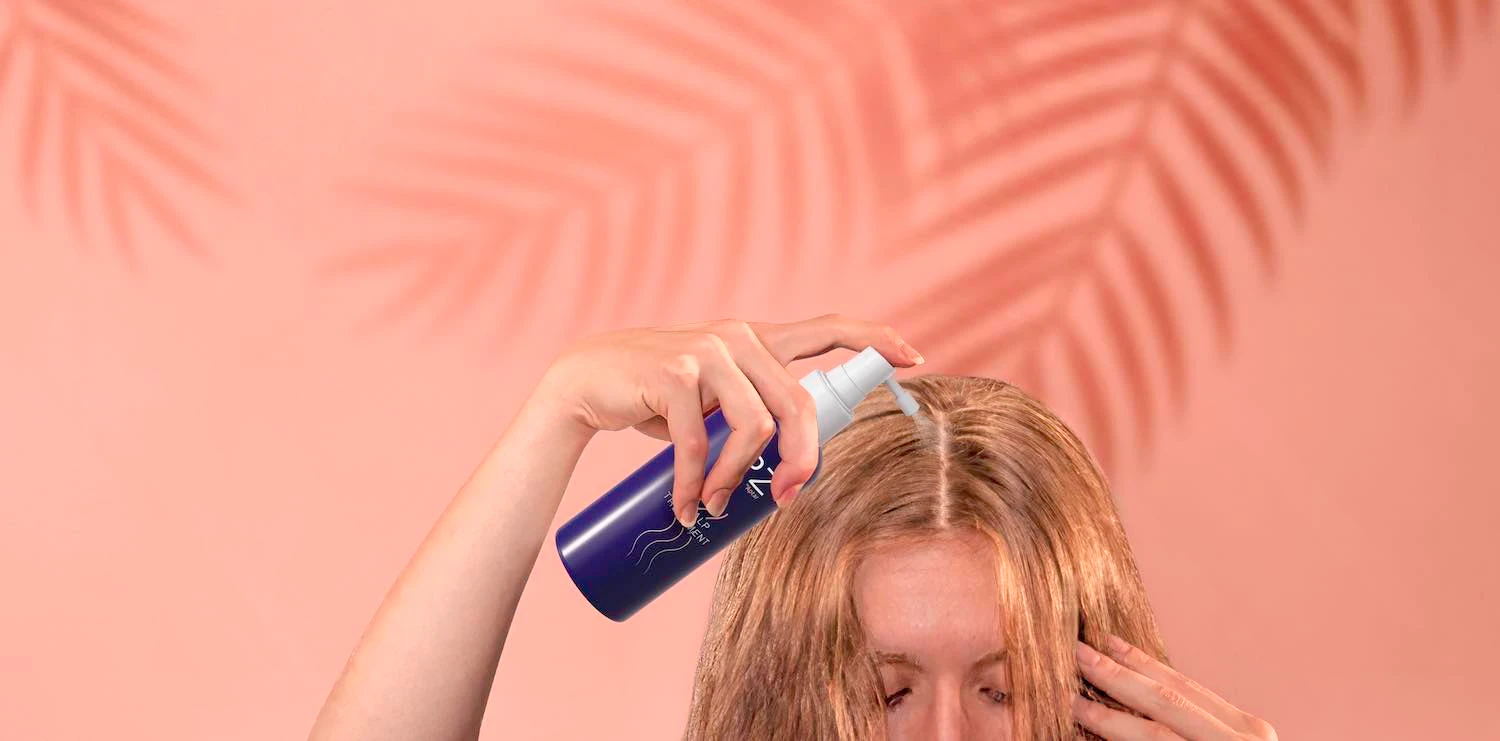 A person sprays a liquid from a blue haircare packaging bottle onto their scalp, parting their blond hair. The background is pink with palm leaf shadows.