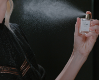 A person in a black lacy top uses a high definition spray fragrance pump, misting perfume from a clear bottle labeled HDS NUNE into the air against a dark background.
