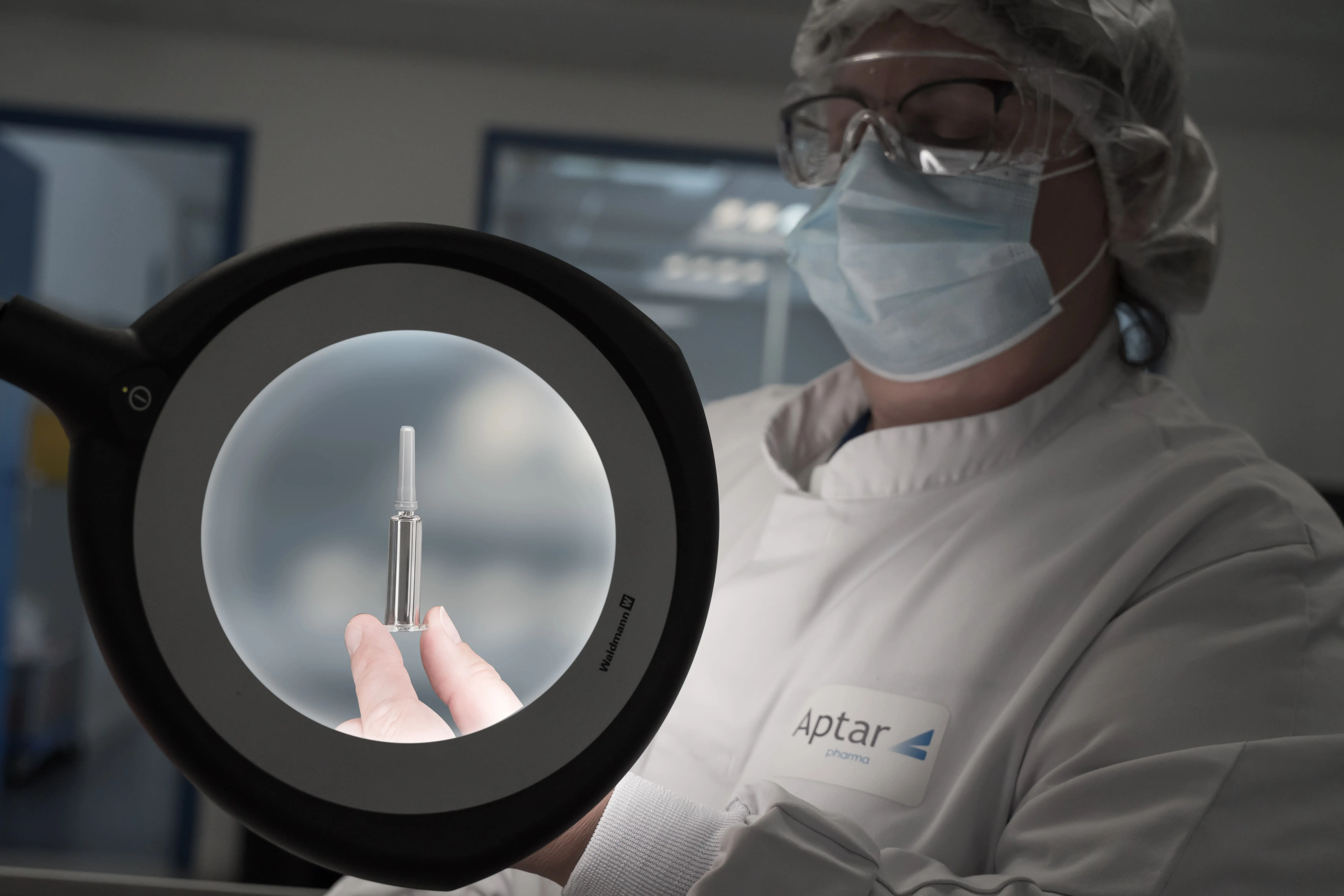 An operator in cleanroom attire is inspecting a component under a magnifying lamp in a pharmaceutical production environment