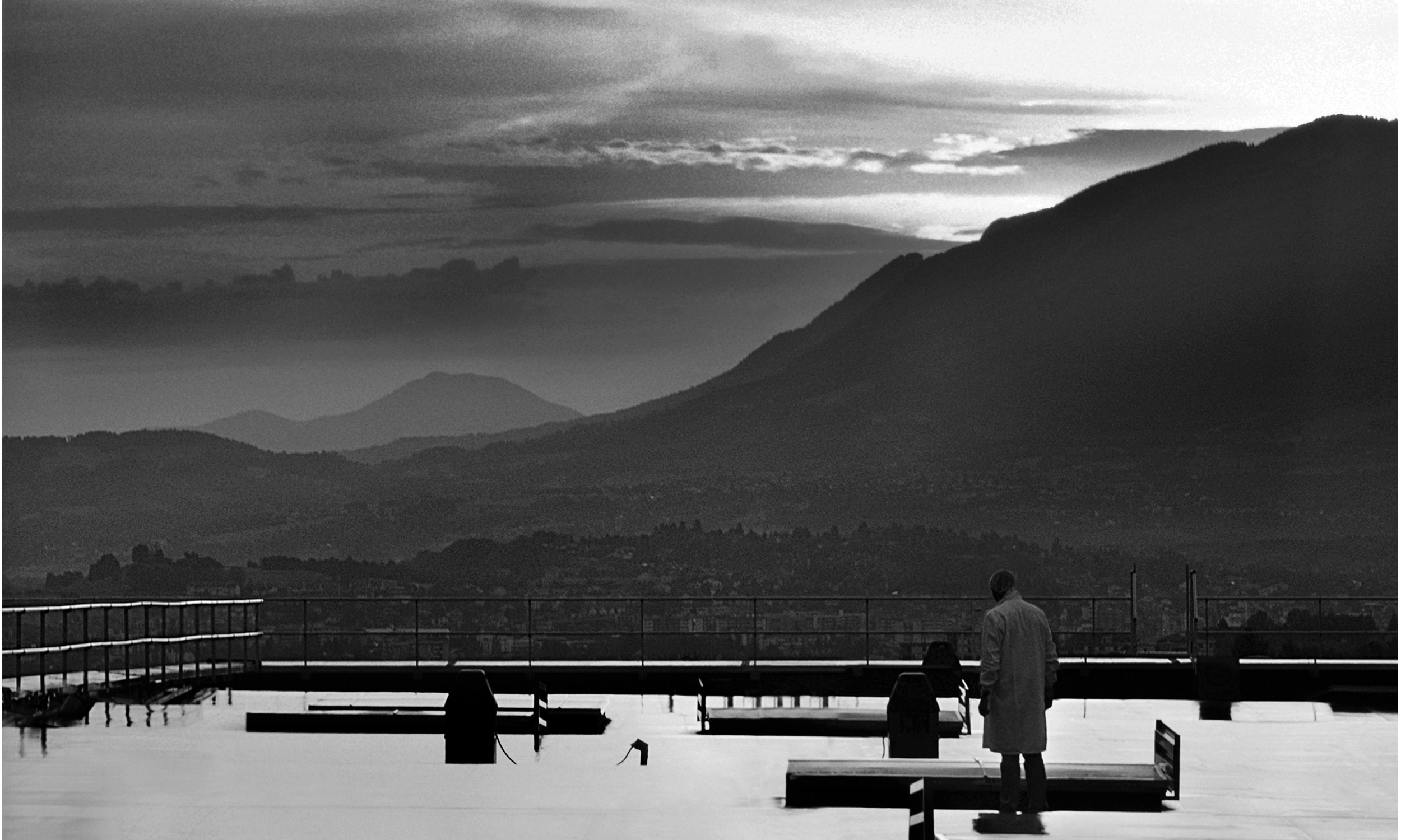 Two people stand on a rooftop at dusk, overlooking rolling hills and mountains beneath a dramatic sky. The black and white image, much like a Non-Guided Lipstick Mechanism labeled Iconic, evokes a moody, contemplative atmosphere.
