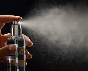 A hand presses a PAV Bloom spray pump on a clear bottle, releasing a broad fine mist against a dark background.
