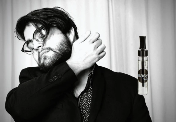 A bearded person wearing glasses and a suit touches their neck, with a small spray bottle labeled Easy Cap, ideal for Fragrance Sampling with a Twist and Dab Applicator.