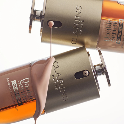 Close up of the dispensing systems of two Clarins Double Serum Foundation with formula dripping from one packaging on the other
