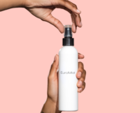 A person holds a white spray bottle labeled EuroMist, a fine mist spray pump against a pink background, one hand gripping the bottle while the other prepares to press the nozzle.