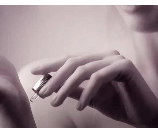 A close-up of a person using a fragrance applicator named Note dropper, applying liquid possibly serum or perfume to their wrist. The image has a soft, muted tone, with only part of the persons face and hands visible.