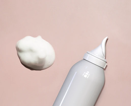 An aerosol actuator for whipped formulas named Aspire with a pointed nozzle dispenses a dollop of white foam against a soft pink background.
